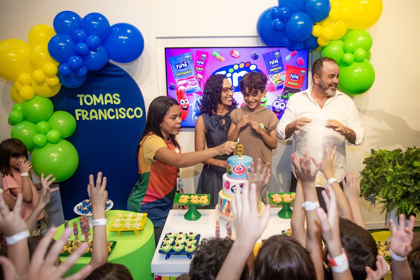 Família cantando parabéns sob arco de balões em festa infantil